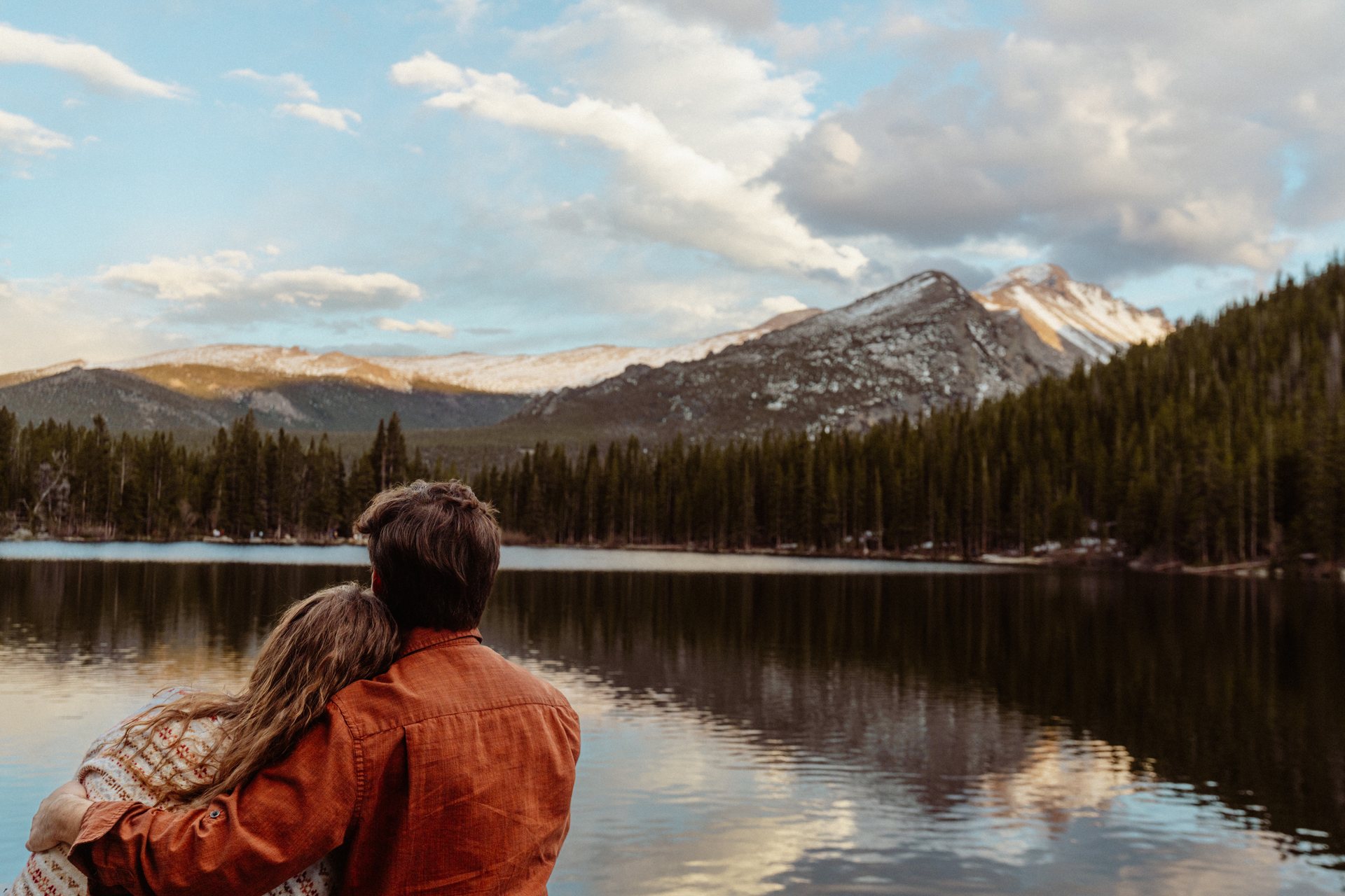 Couple seated on lakeside rocks below a snow-capped peak, engagement session in Rocky Mountain National Park