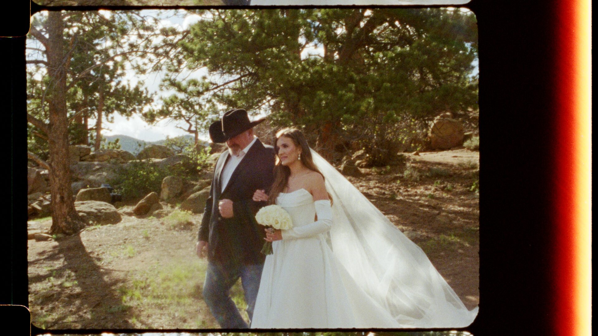 16mm film still — Emma and Ryan walking arm-in-arm, bride's veil flowing, Rocky Mountain granite boulders and pines in the background. The fine grain structure of 16mm is visible at close inspection.