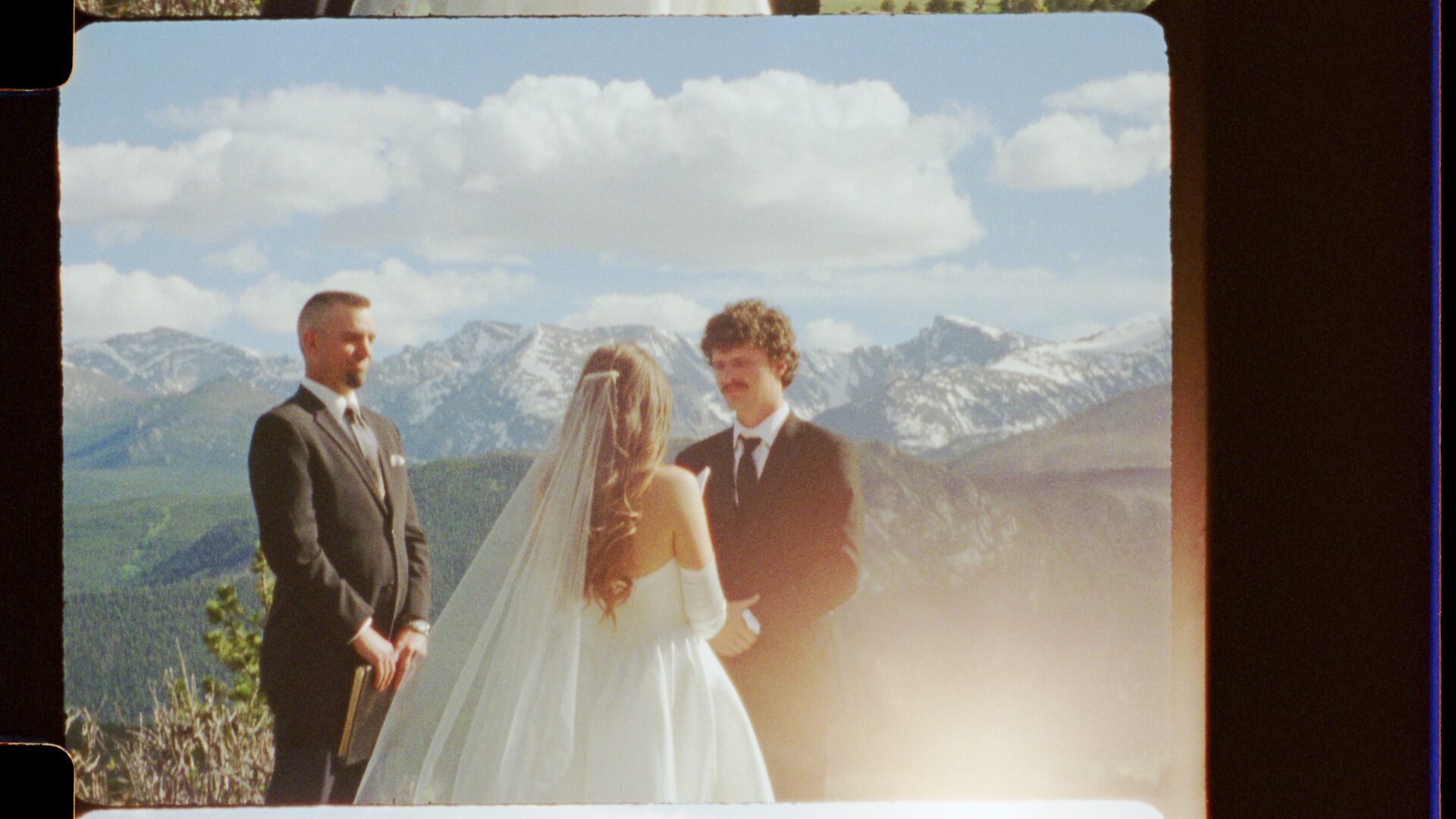 16mm film still — Emma and Ryan's elopement ceremony, wide shot with officiant, snow-capped Rocky Mountains in the background under bright blue sky.