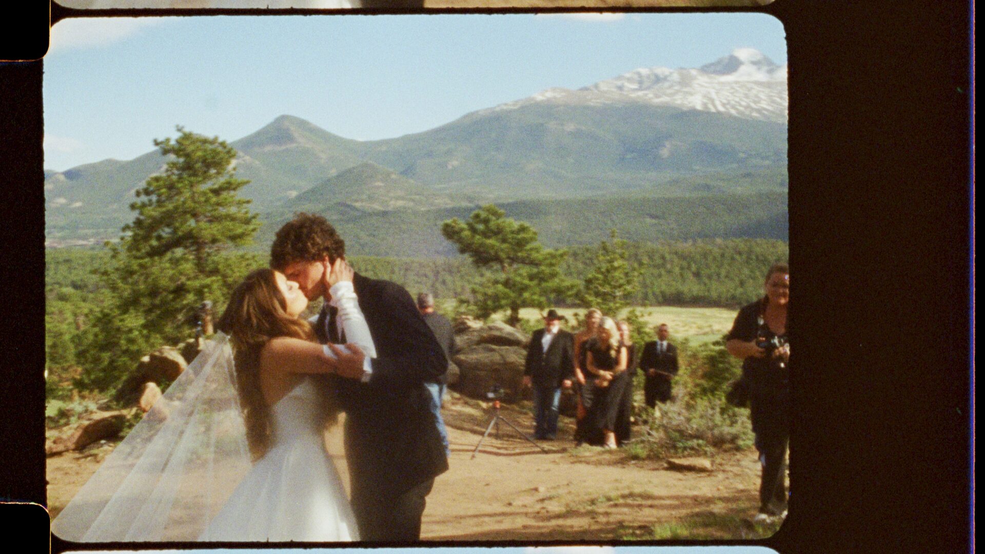 16mm film still — Emma and Ryan's first kiss at their Rocky Mountain elopement, guests and snow-capped peaks visible in the background. Shot on 16mm cinema film by Motus Weddings.