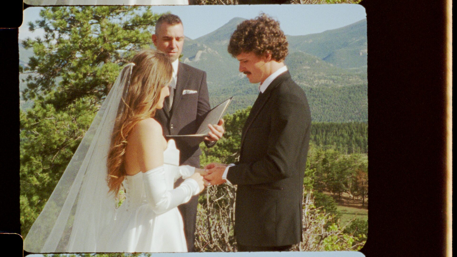 16mm film still — Emma and Ryan exchanging rings during their elopement ceremony, officiant reading, forested Rocky Mountain backdrop.
