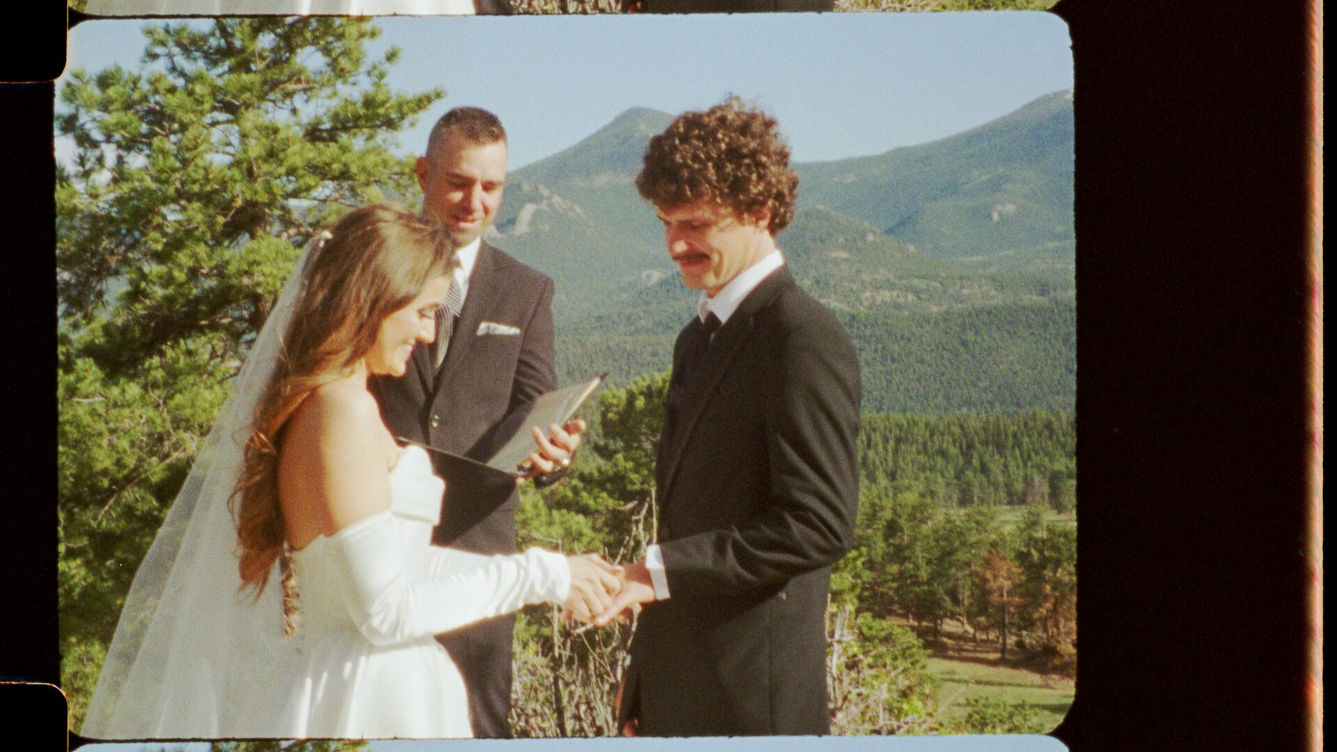 16mm film still — Emma and Ryan exchanging vows, both smiling, Rocky Mountain forest and peaks behind them. The joy of the moment is unmistakable even in a film still.
