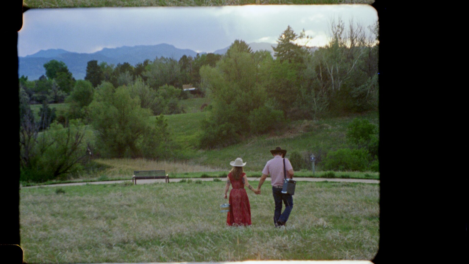 16mm film still — Kesia and Brian walking together through a lush Colorado field, Rocky Mountains visible in the distance, black film borders visible on frame edges.