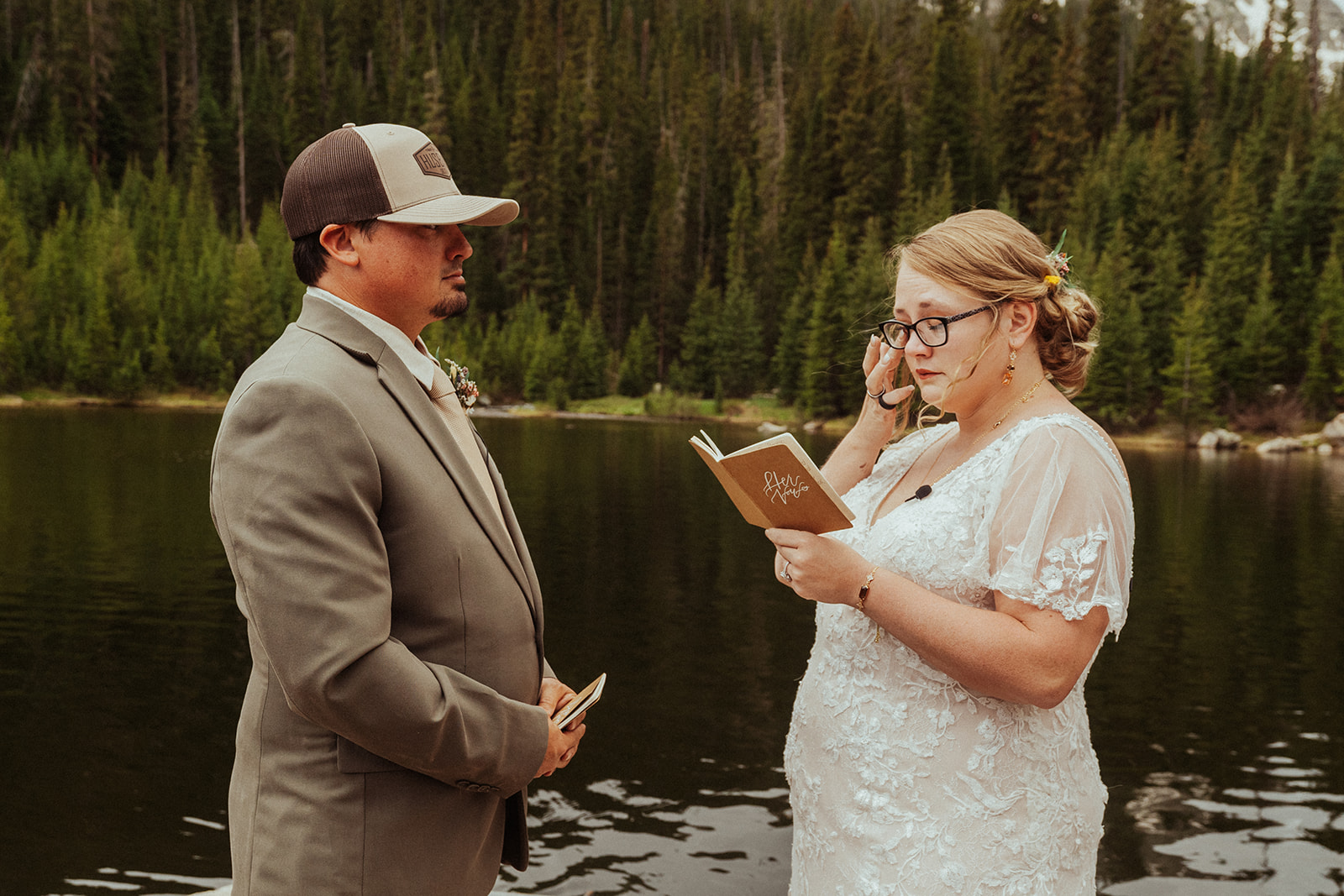 Bride reading her vows in front of a small lake with groom listening, vows in hand. Bride is crying and touching, wiping tears away from her eye in an elopement.
