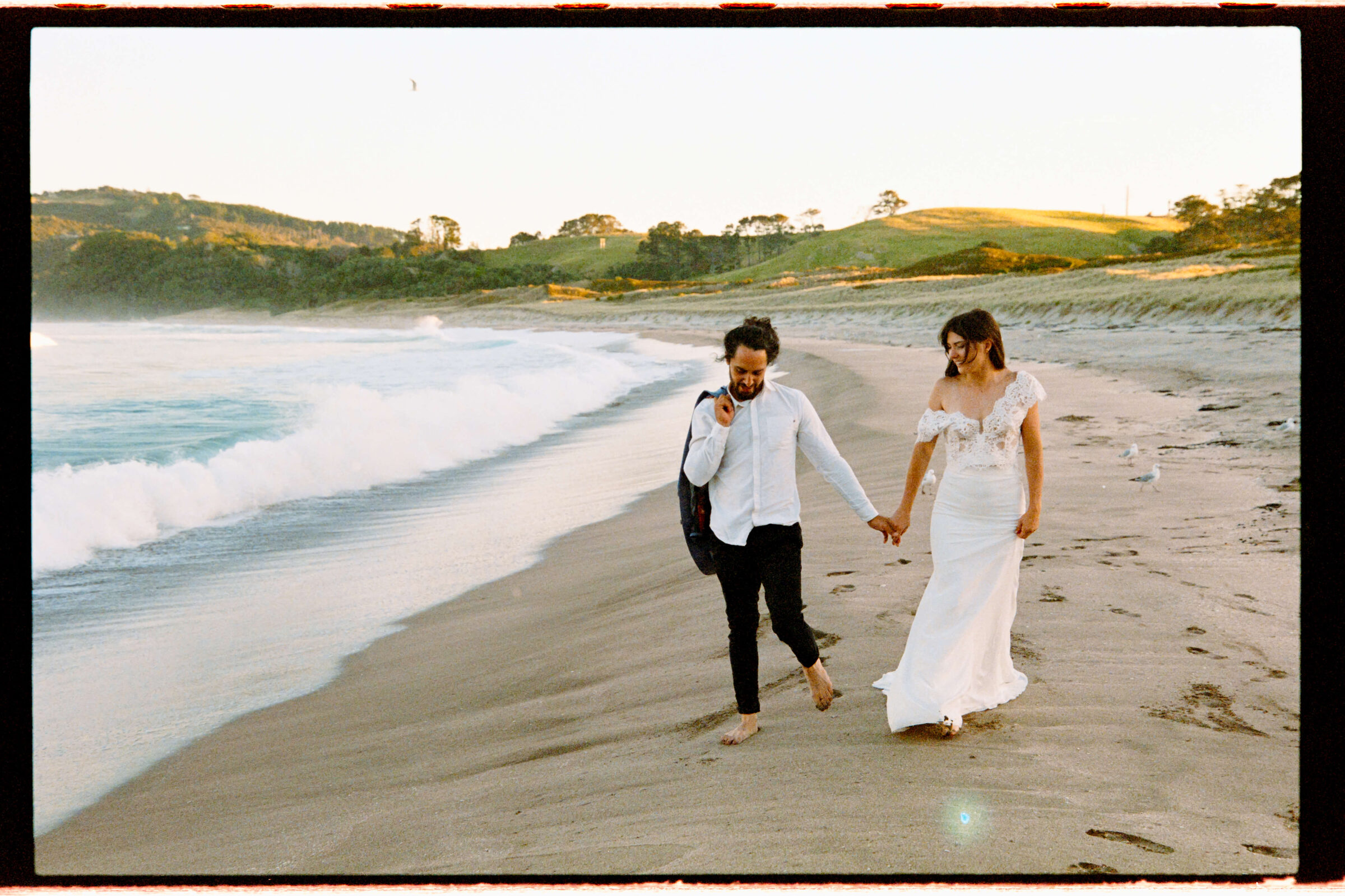 Couple walking down the beach holding hands. Groom with jacket over his right shoulder. Bride smiling down at him. The right dress strap is resting down on her off of her shoulder