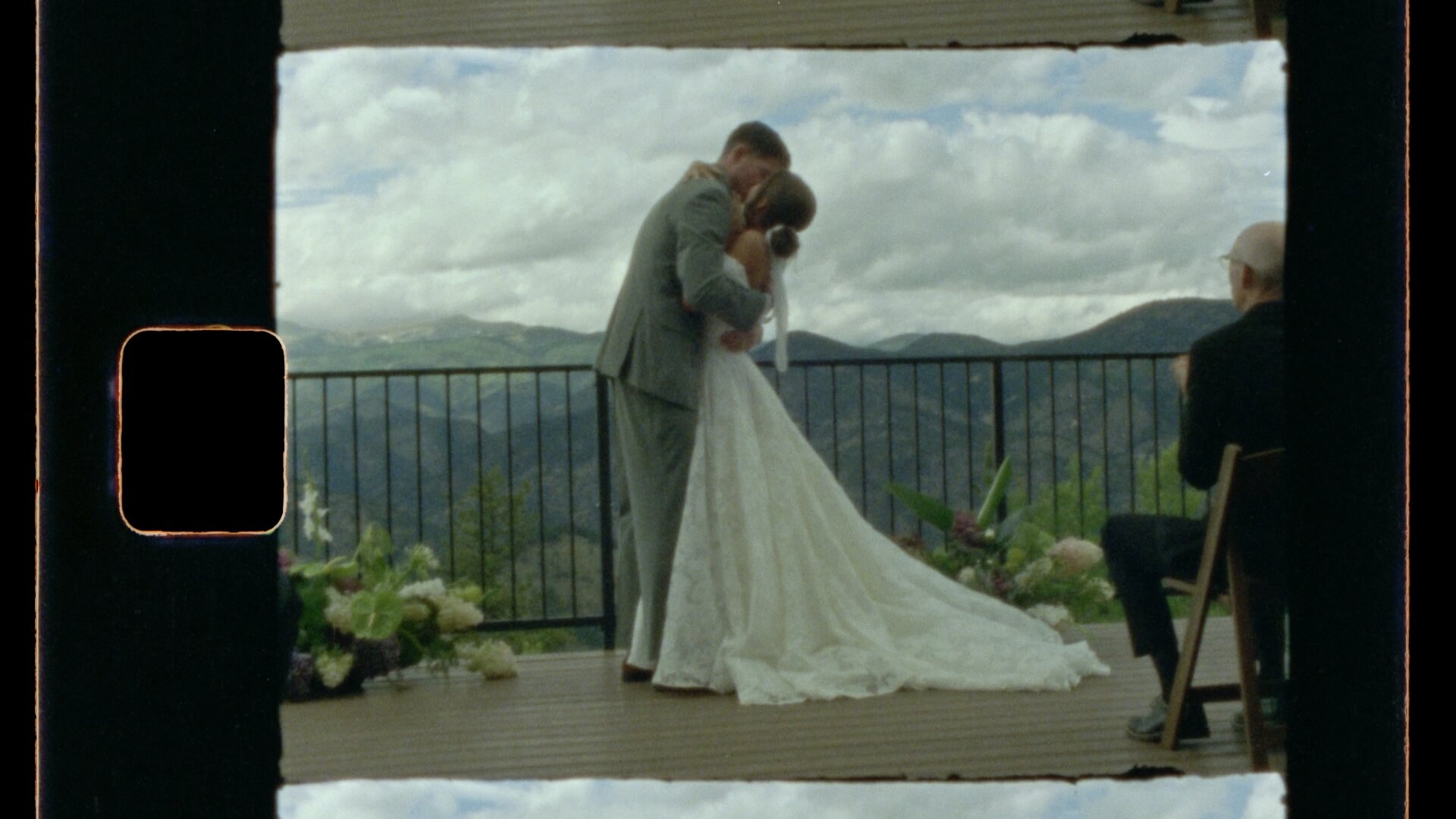 Super 8mm wedding film frame — couple sharing their first kiss on a mountain deck ceremony, Colorado peaks stretching across the horizon. Organic film grain, Kodak color science, and the characteristic film frame border of Super 8mm telecine.