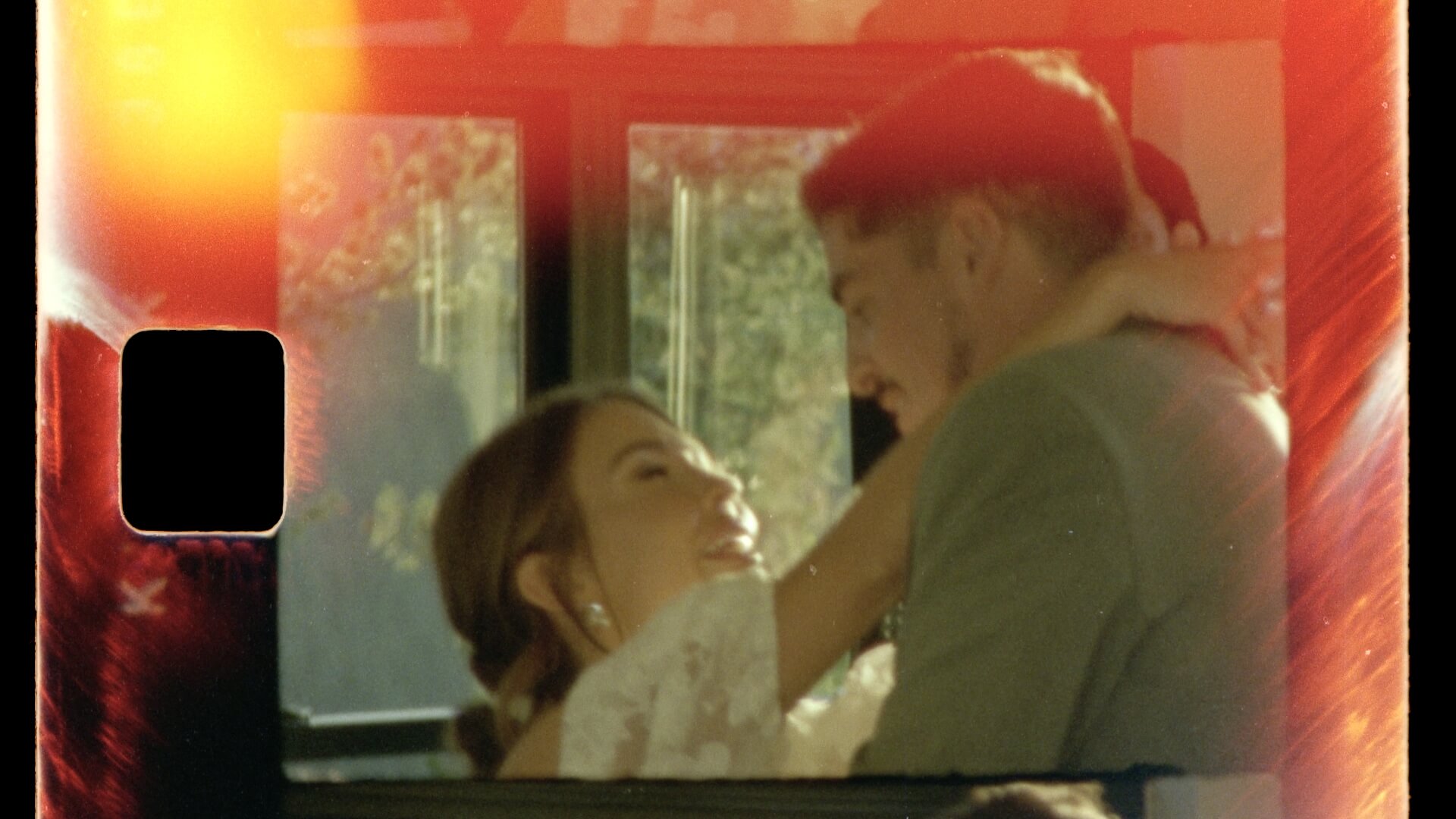 Super 8mm wedding film frame — intimate indoor moment, couple close, warm orange light leak spreading across the frame. The photochemical warmth and organic color rendering of Kodak Super 8mm film stock.